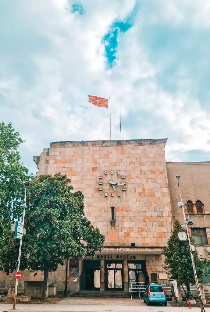 Front of the old Skopje railway station, now a museum, with a broken clock stopped at the moment of the 1963 earthquake and the Macedonian flag flying above.