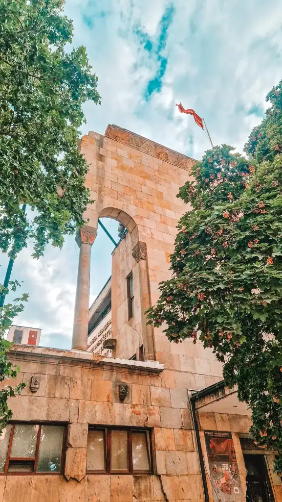 Exterior view of the Museum of the Macedonian Struggle in Skopje, featuring a stone archway with classical columns and the Macedonian flag flying above, framed by leafy trees.