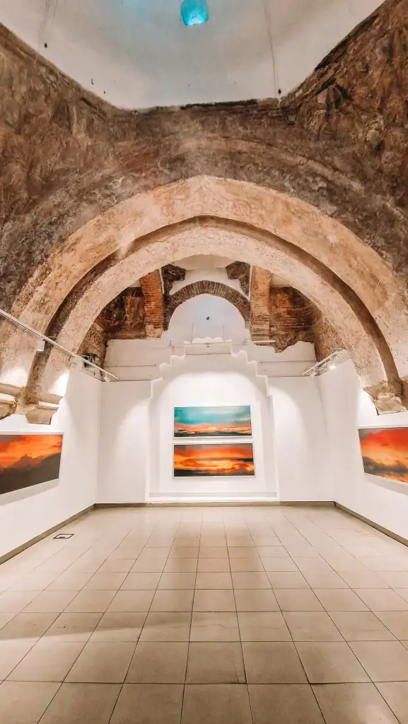 Art gallery inside a restored Ottoman building in Skopje, with arched brick ceilings and landscape photos of vivid sunset skies mounted on white walls.