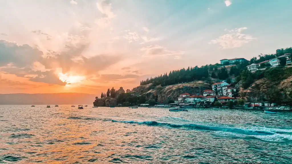 The sun sets over Lake Ohrid, casting warm light over the iconic cliffside church and boats gently bobbing on the water, a serene highlight of North Macedonia.
