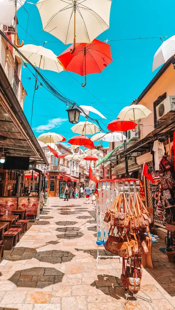 A cobbled pedestrian street in the Old Bazaar lined with artisan stalls and souvenir shops, with red and white umbrellas suspended overhead casting shadows on the ground.