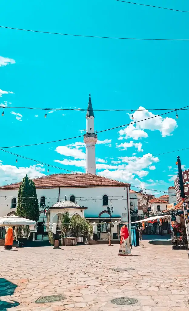 A central square in Skopje’s Old Bazaar, showcasing a white mosque with a tall minaret, string lights above, and stalls selling souvenirs and clothing.