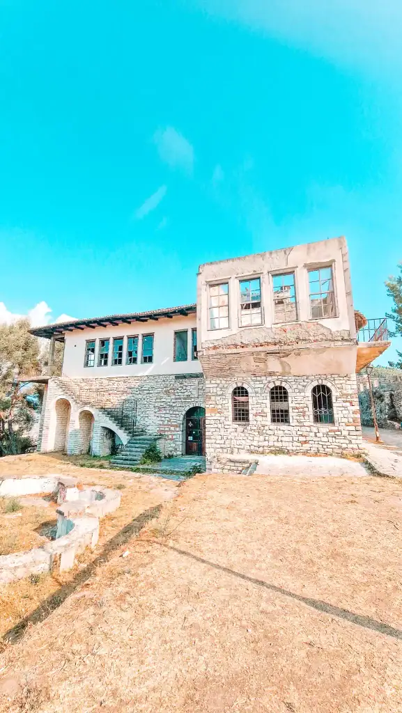 Weathered stone building with arched doorways and a partially restored upper level inside Berat Castle, surrounded by dry grass and ruins.