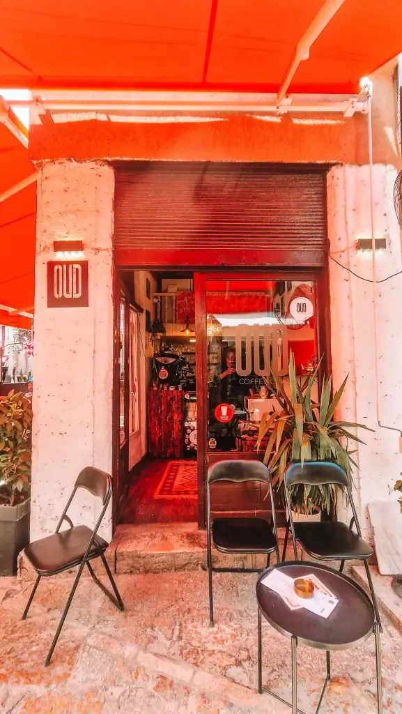 The entrance to OUD Coffee Club in Skopje’s Old Bazaar, with two black chairs and a small table outside under a red canopy, and a rustic wooden interior visible through the glass door.