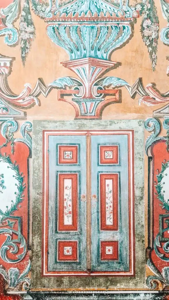 A close-up of an ornately painted double door in a mosque, surrounded by intricate floral and scrollwork details in turquoise, red, and gold. The faded texture adds to its historic charm.