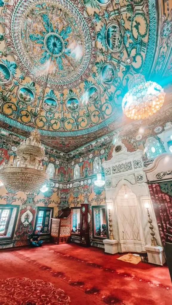 Inside the Painted Mosque, intricate ceiling art and Arabic calligraphy cover every surface. Multiple chandeliers hang from the dome, and the rich red carpet fills the prayer area.