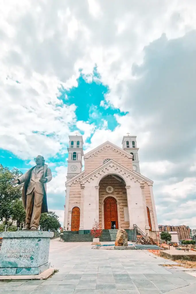 A large statue of Ibrahim Rugova stands in front of the grand white stone Cathedral of Saint Mother Teresa in Pristina, Kosovo, under a partly cloudy sky.