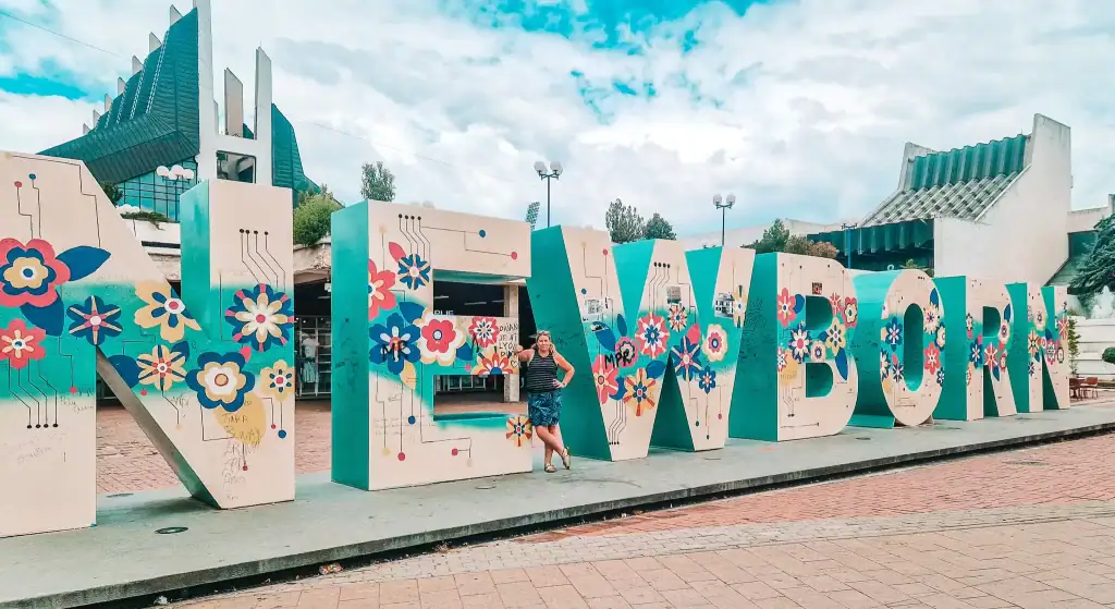 Em poses with the colorful “NEWBORN” monument in Pristina, covered in flower-themed street art. The National Library’s unique architecture rises behind the installation.