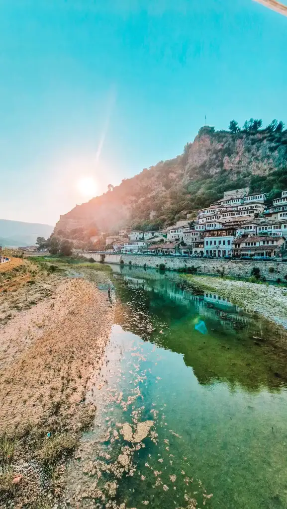Warm sunlight setting behind a rocky hill lined with Ottoman houses in Berat, reflected in the calm waters of the Osum River.