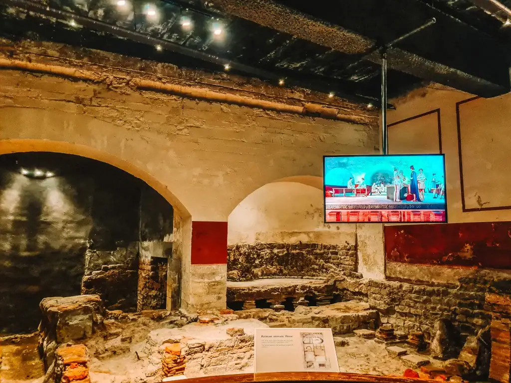Interior ruins at the Roman Baths showing exposed stone foundations, with a digital screen overhead displaying an animated reconstruction of the ancient space.