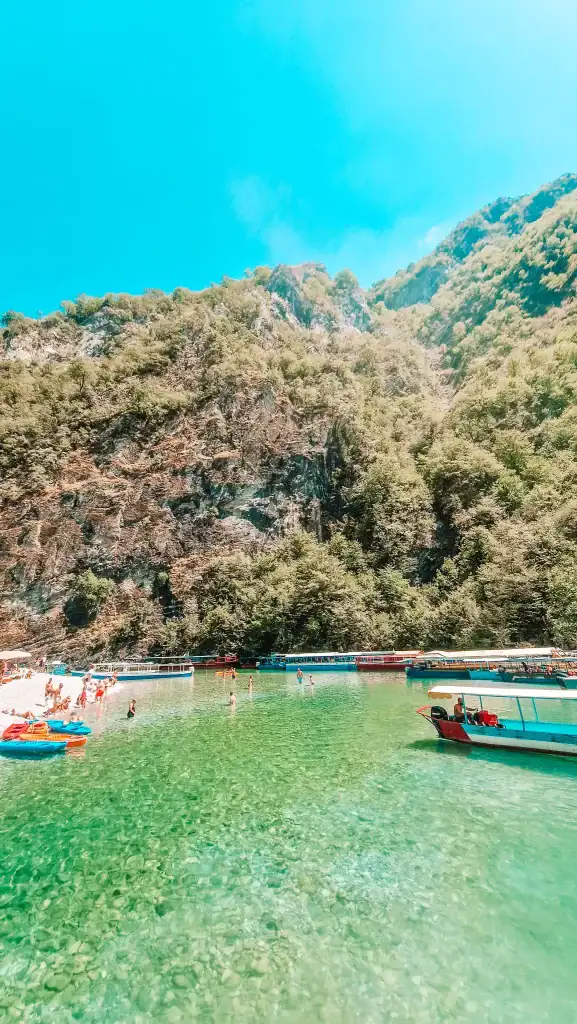 Swimmers wade through strikingly clear green water near colourful boats and a small pebbled beach, surrounded by steep forested cliffs in the Albanian Alps.