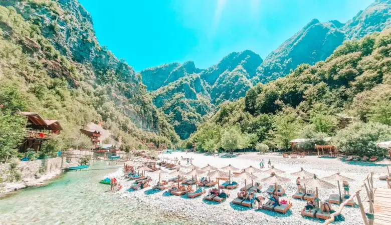 Tourists relax on wooden sunbeds under straw umbrellas beside a rocky riverbank, surrounded by dense forest and mountain peaks near Shkoder.