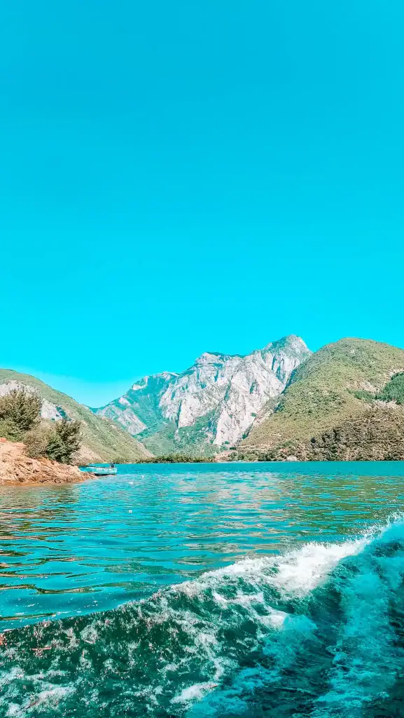 Waves trail behind a boat gliding across Komani Lake, with dramatic craggy peaks rising in the distance under a cloudless blue sky.