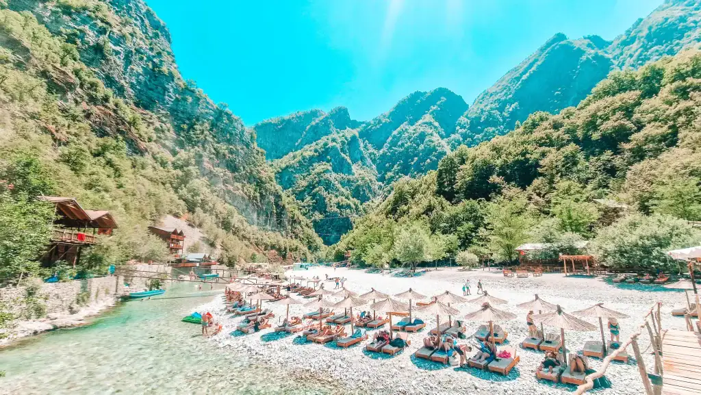 Sun loungers and straw umbrellas line the rocky riverbank at the Shala River in Albania, surrounded by steep green mountains and wooden cabins, offering a hidden escape for nature lovers.