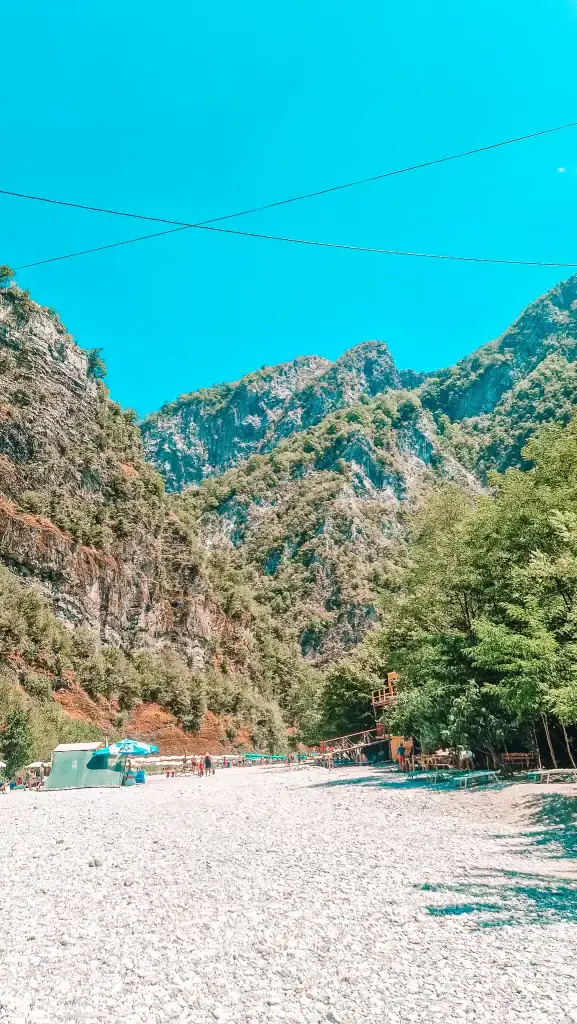 Wide stony beach lined with sun umbrellas and lounge chairs at the base of towering green mountains, a popular local escape just outside Shkoder.