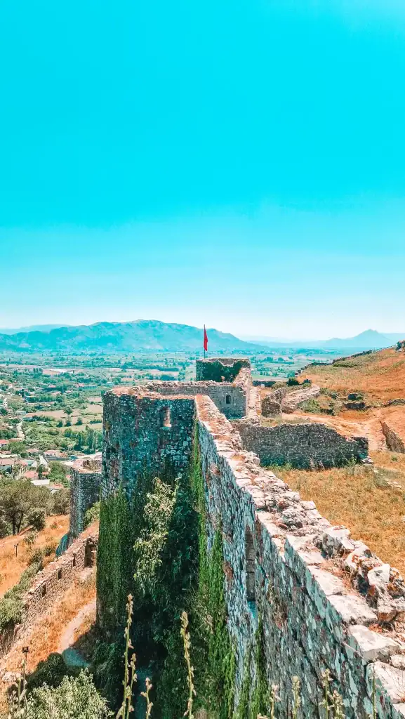 The stone walls of Rozafa Castle stretch across a hilltop overlooking the Albanian countryside, marked by a red flag and sweeping views of the Shkodër valley below.
