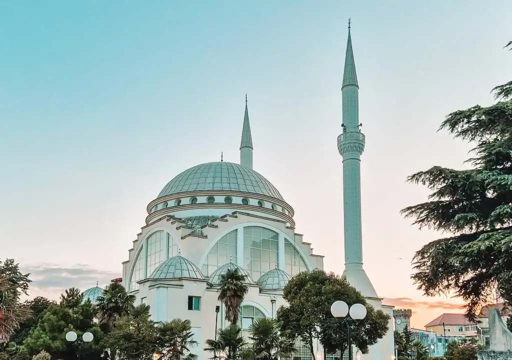 A grand white mosque with a central dome and two tall minarets framed by palm trees, photographed at sunset against a pastel sky.