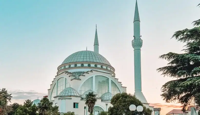 A grand white mosque with a large central dome and two slender minarets rises against the soft hues of a sunset sky. Surrounded by palm trees, the mosque is a peaceful landmark in a city served by Albania buses.