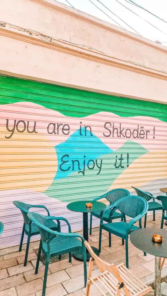 Outdoor café seating in front of a painted shutter with bold, colourful shapes and cheerful text welcoming visitors to Shkoder.