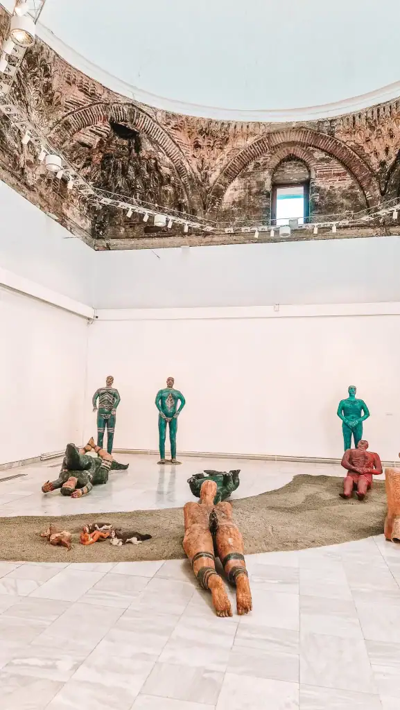 Contemporary art installation of abstract human figures in vivid colors arranged across a marble floor under a dome with historic brick and fresco details.