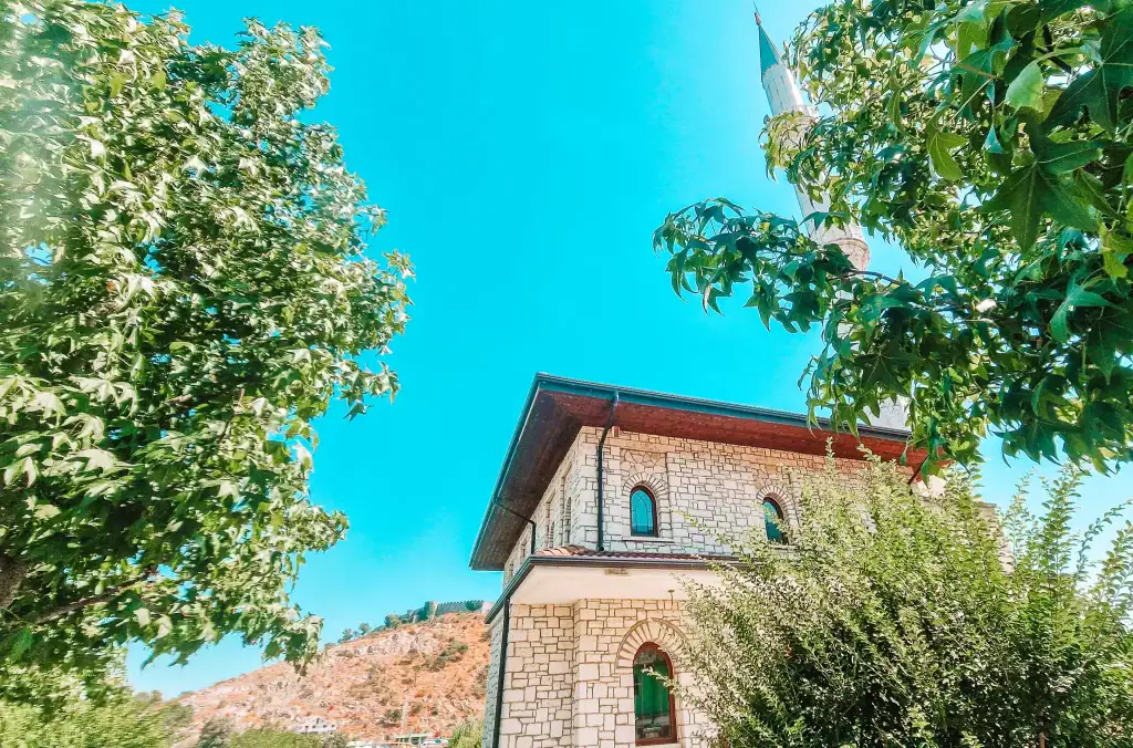 A quaint stone mosque with arched windows and a tall minaret peeks through leafy trees, with the walls of Rozafa Castle perched on a hill behind.