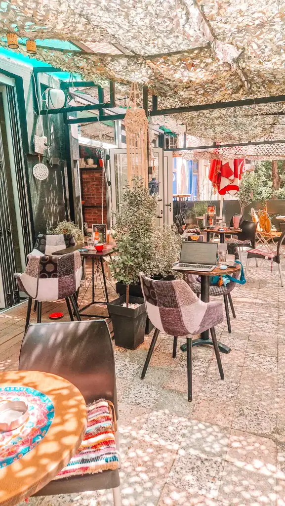 A shaded outdoor area of a Skopje cafe decorated with patchwork chairs, hanging macramé, and lush potted plants. Sunlight filters through a camo-style canopy, and a laptop sits open on one of the tables.