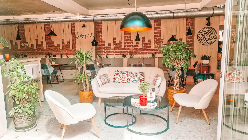 A cosy and modern Skopje cafe interior with light wood and brick walls, leafy plants, and a central seating area featuring a white couch with floral cushions and a set of round glass tables.