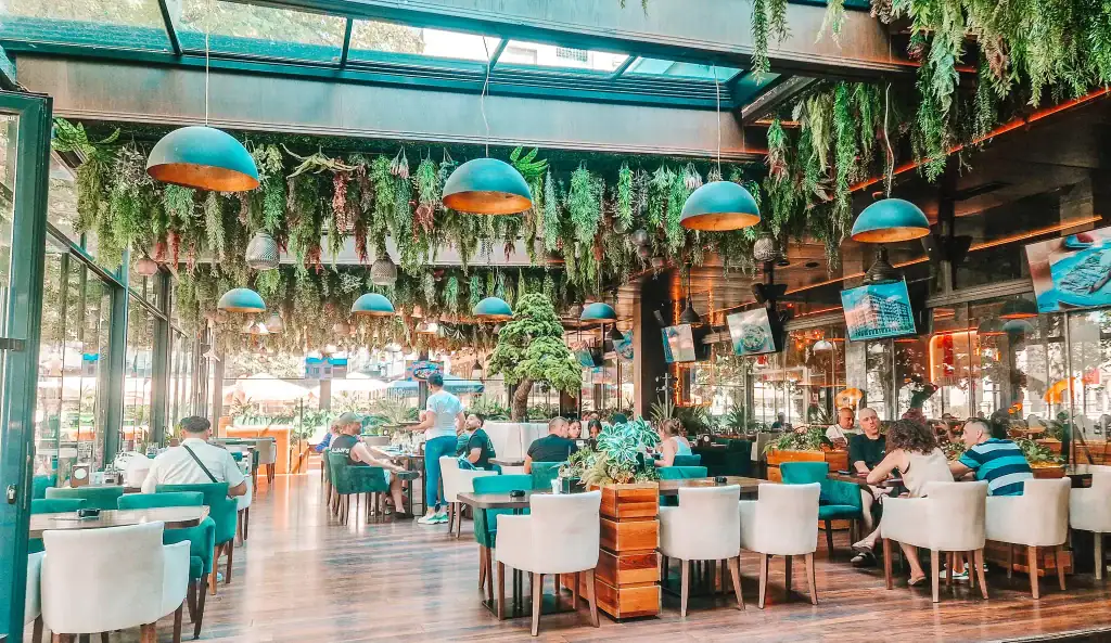A spacious, vibrant cafe in Skopje filled with hanging greenery, teal and cream chairs, and floor-to-ceiling windows. Groups of people relax under dome pendant lights in a lush indoor garden-like setting.