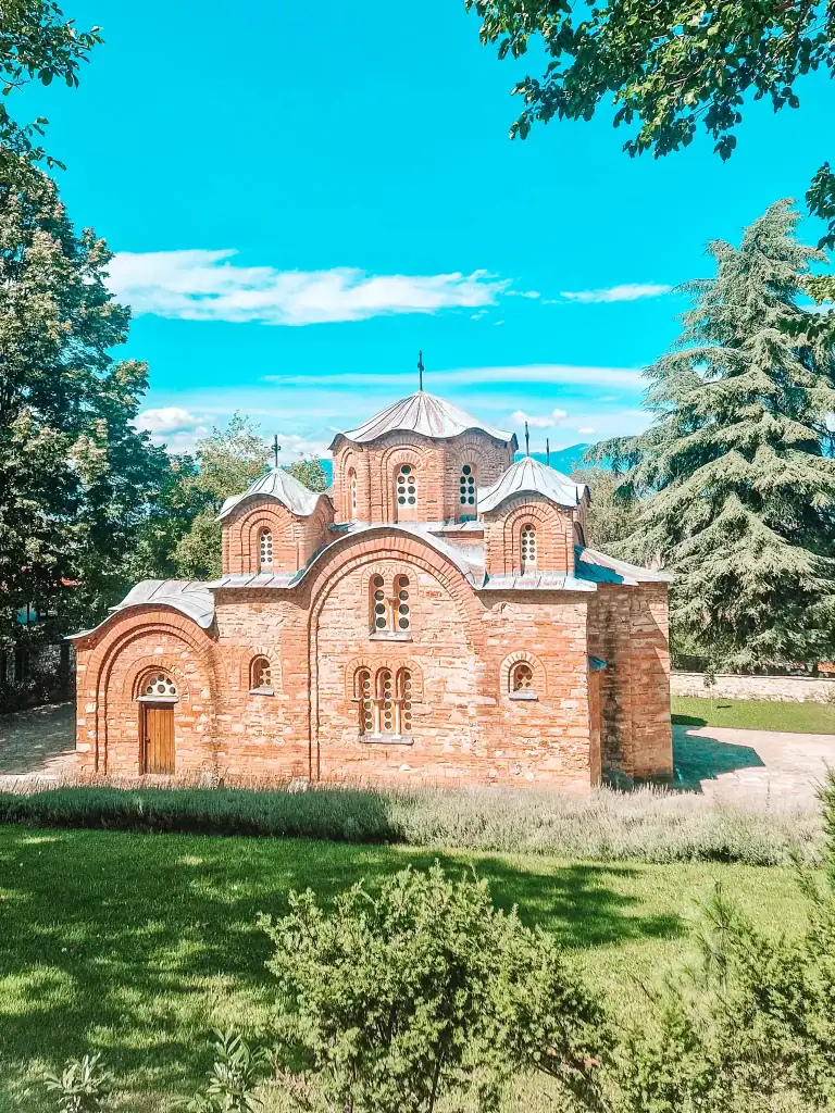 The red-brick exterior of the Church of St Panteleimon in Gorno Nerezi, surrounded by manicured greenery and tall trees under a bright blue sky.