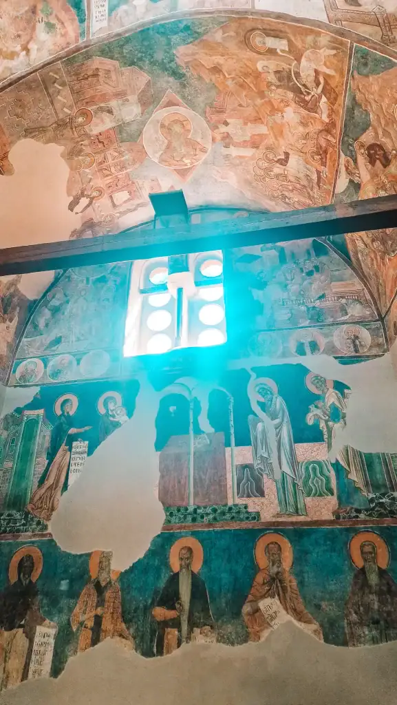 Wall frescoes inside a centuries-old church show rows of haloed saints, partially faded with time, beneath a glowing window with circular panes.