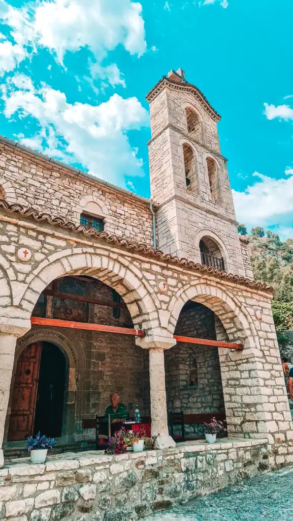Tall stone church tower with arched windows and a tiled roof, part of a historic church with an arched portico in Berat.