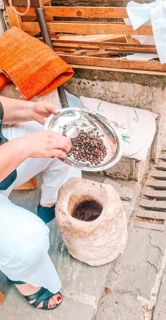 A woman holds a tray of roasted coffee beans above a stone mortar, demonstrating a traditional coffee preparation method in a cultural craft setting.