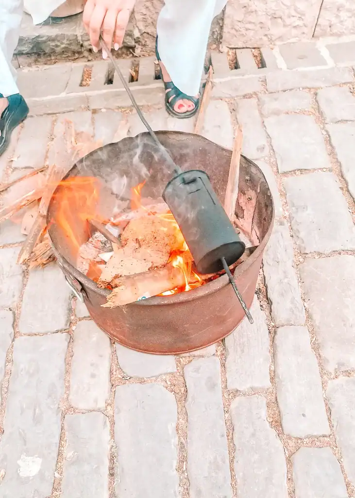 A hand stirs a traditional coffee roaster over an open flame in a metal pot on cobblestone pavement in Gjirokaster. The fire crackles with wood and cardboard, part of a street-side coffee preparation method.
