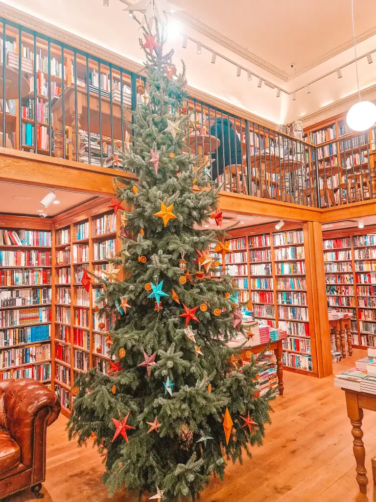 Tall Christmas tree decorated with colorful paper stars and dried orange slices inside Topping & Co, surrounded by wooden bookshelves stacked with books.