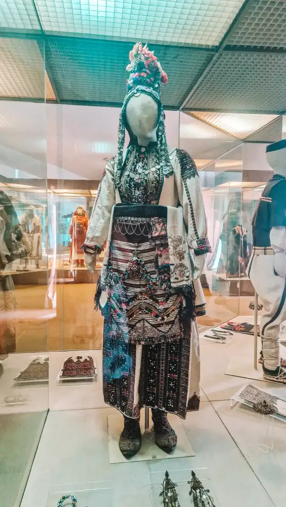 Traditional Macedonian bridal costume on display in a glass case, richly embroidered with colourful beads, metal elements, and layered textiles.