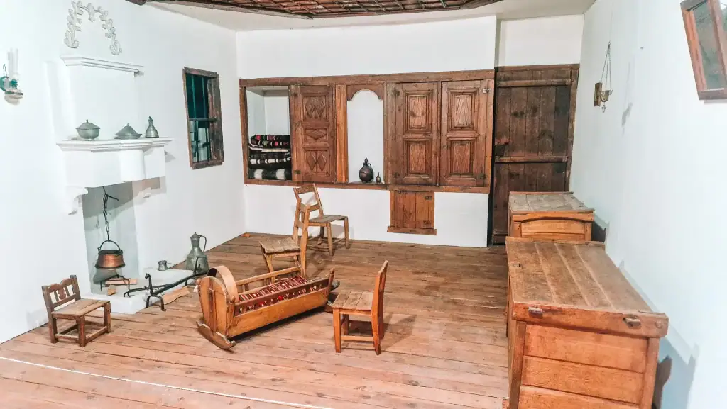 Museum exhibit recreating a traditional Macedonian home interior with wooden furniture including a cradle, chairs, and storage chests, along with a white fireplace and woven blankets neatly stored in a wall alcove.