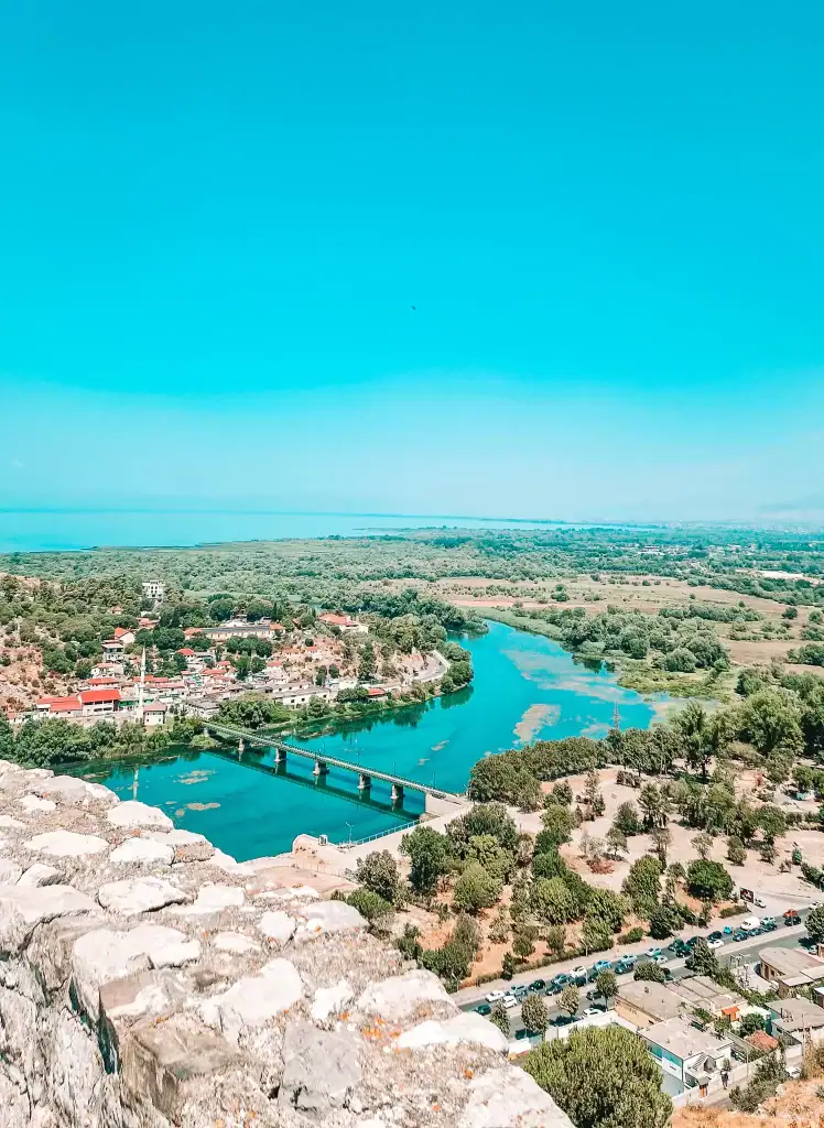 Looking down from the castle walls, the image shows the winding Buna River with a bridge connecting two sides of the town, surrounded by greenery and small red-roofed buildings.