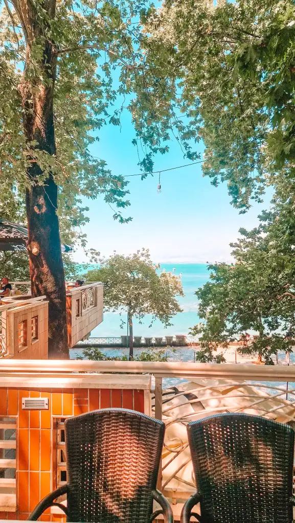 Outdoor seating at a terrace café shaded by leafy trees, overlooking the blue waters of Lake Shkodra on a peaceful sunny afternoon.