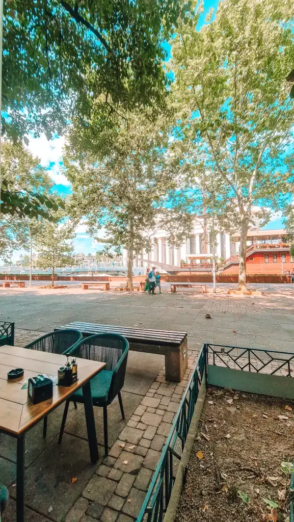 A riverside cafe terrace in Skopje shaded by tall trees with a view of neoclassical buildings and a red boat-style restaurant across the water. A wooden table and two chairs are placed on a stone patio in the foreground.