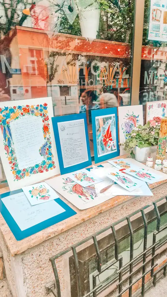 A window display outside a Skopje cafe featuring colorful hand-painted art, calligraphy, and flowers arranged on a stone ledge with reflections of the street scene behind it.
