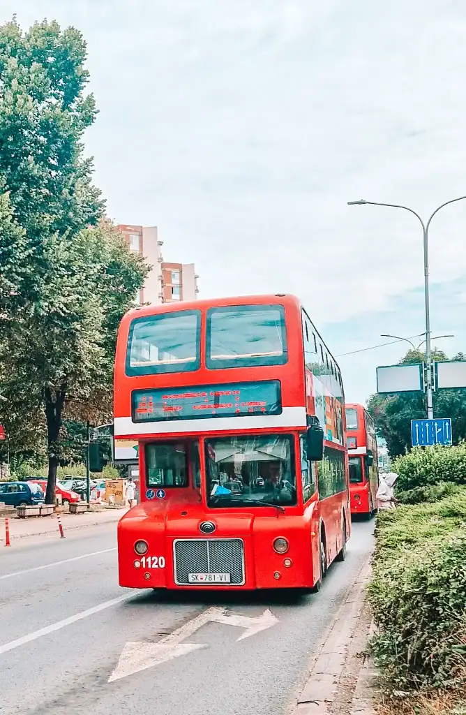 A bright red double-decker bus with a digital route display travels down a city street in Skopje, flanked by trees and modern apartment buildings in the background.