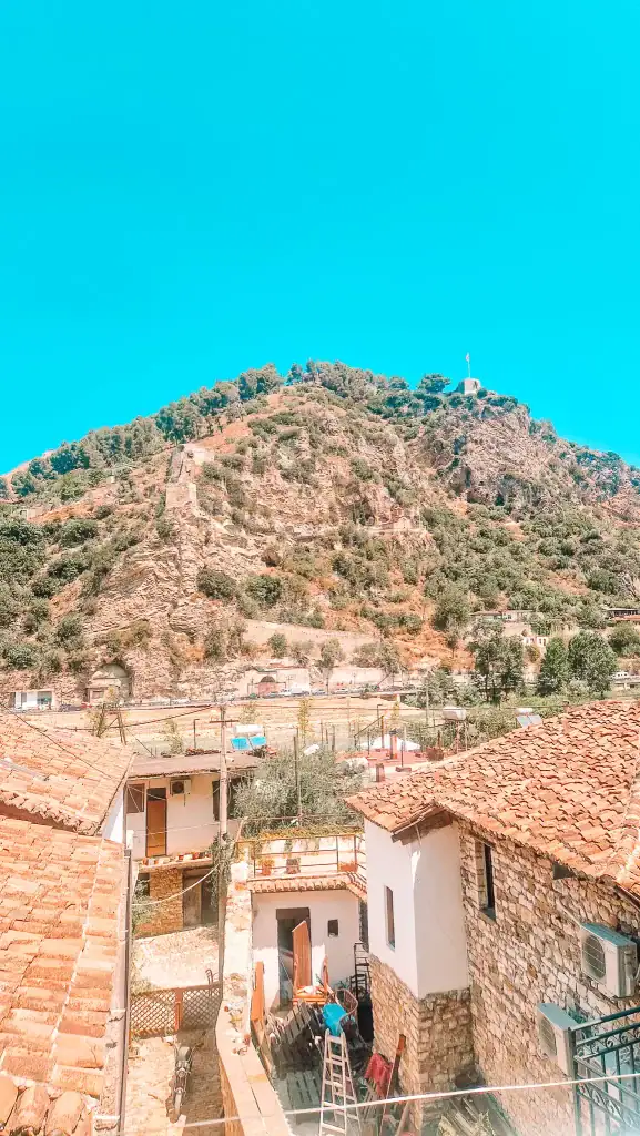 Red-tiled rooftops of stone houses in the foreground with a steep rocky hill topped by a flag in Berat.