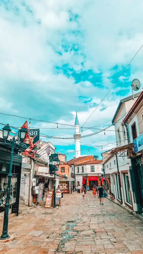 Bustling street in Skopje’s Old Bazaar featuring shops, hanging lights, and a tall mosque minaret rising above the historic cobblestone lane.