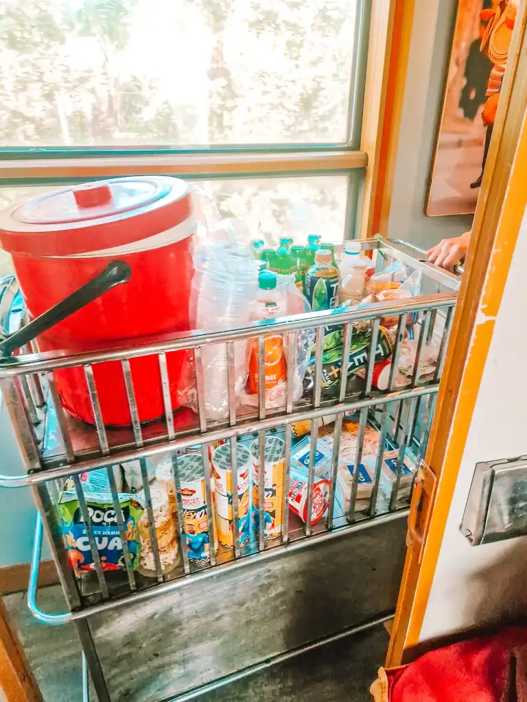 A rolling metal snack cart on a Vietnamese train is packed with bottled drinks, instant noodles, and snacks like popcorn and chips. A red cooler on top hints at hot beverages being served, including Vietnamese coffee options.