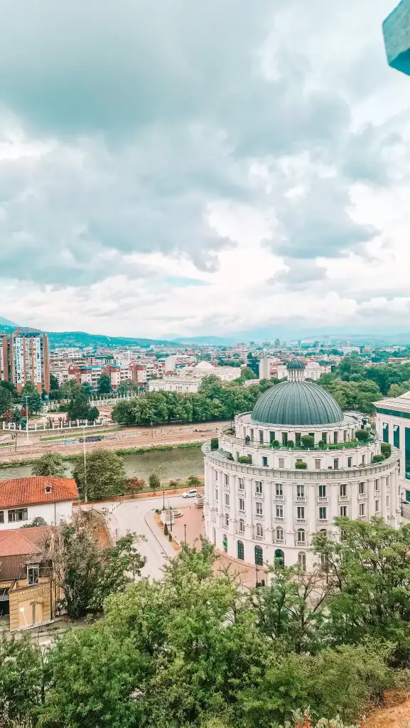 A cloudy panoramic view over central Skopje with a domed neoclassical building in the foreground, the Vardar River winding through the city, and clusters of modern and Soviet-era buildings stretching into the distance. This wide-angle shot adds context to any Skopje guide by showing the city’s blend of architecture and natural surroundings.