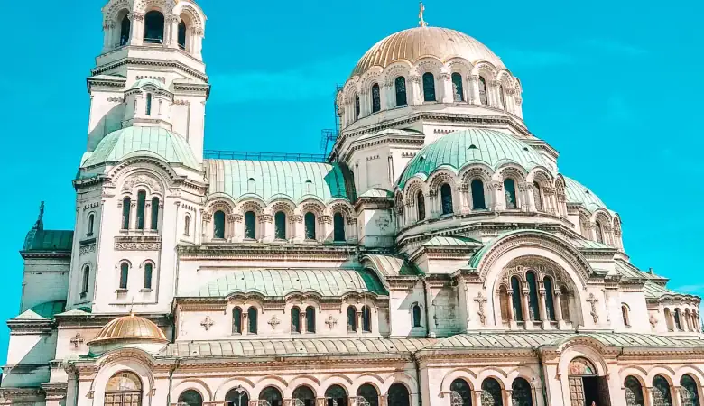A wide-angle shot of the Alexander Nevsky Cathedral in Sofia, showcasing its intricate Neo-Byzantine architecture and green domes glowing under clear daylight.