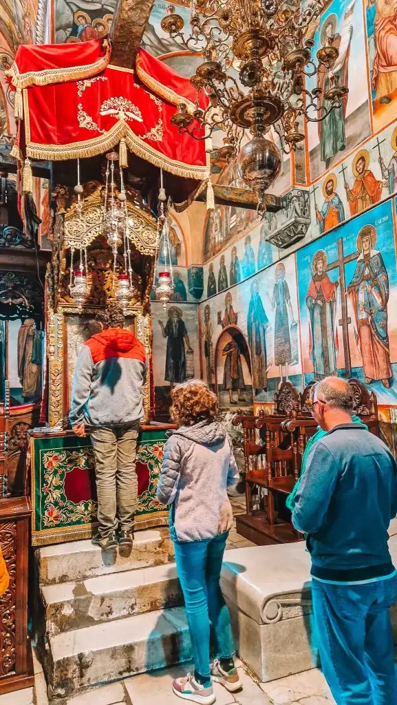 People praying and observing a decorated religious shrine inside a richly painted Orthodox church with colourful frescoes of saints and biblical scenes covering the walls.