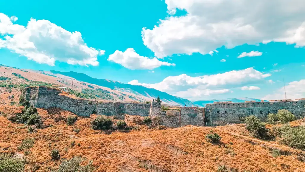 Ancient stone walls of Gjirokastër Castle stretch across dry, orange hills under a bright turquoise sky, with the Albanian mountains rising in the background. A few scattered trees grow along the rugged terrain.
