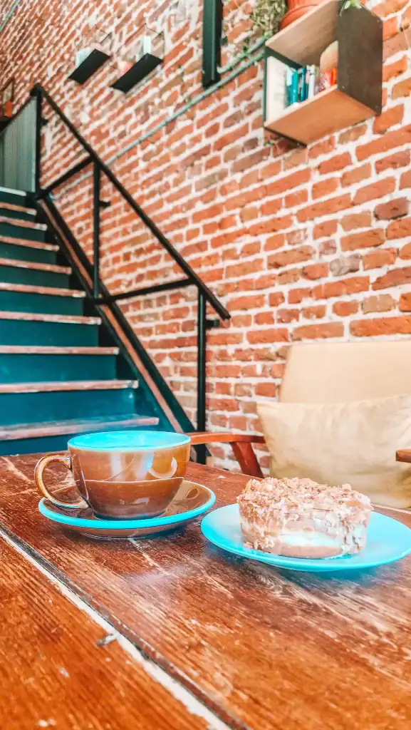 A cosy indoor café corner with exposed brick walls, a wooden staircase, and a rustic table holding a cappuccino in a brown cup with a saucer and a dessert that looks like a nut-covered doughnut. The relaxed setting hints at a quiet spot for coffee lovers.
