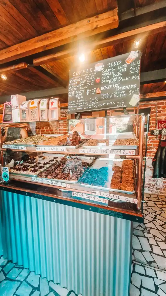 A vibrant café counter with a chalkboard menu listing espresso, cappuccino, teas, and homemade lemonade above a pastry display filled with treats like vanilla glazed, pistachio cream, and Boston cream doughnuts. A great stop for sweet tooths exploring the best cafes in Plovdiv.